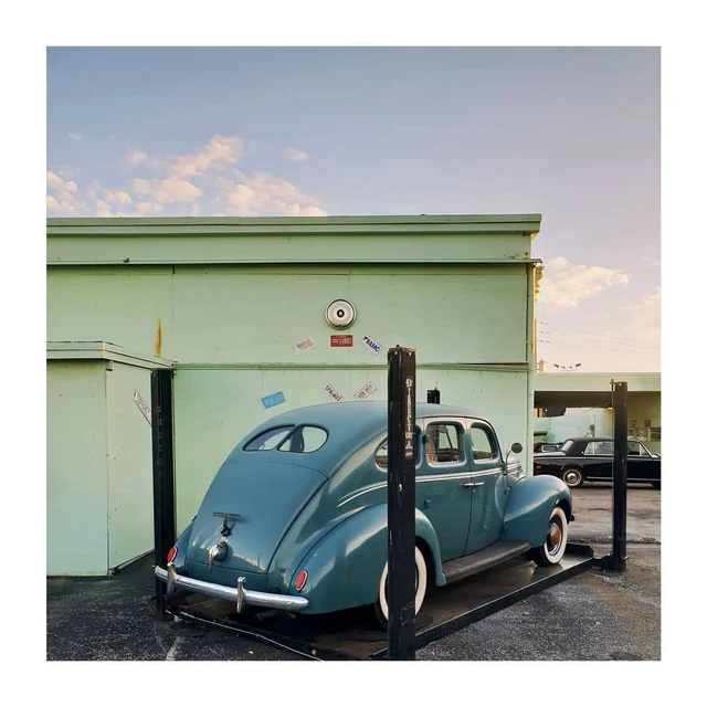 Une voiture ancienne bleue sur un pont élévateur devant la façade vert pâle d’un garage.