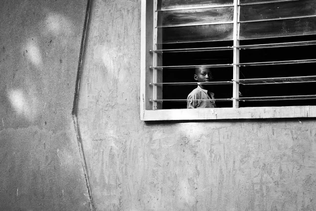 Un enfant regarde dehors derrière les barreaux horizontaux d’une fenêtre, dans un mur éclairé par des taches de lumière, en noir et blanc.