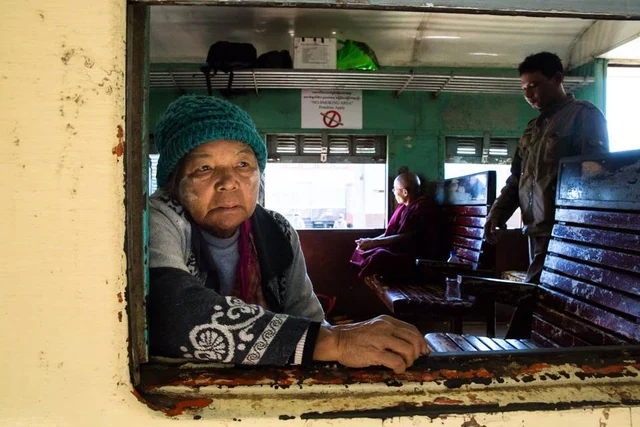 Une passagère portant un bonnet turquoise s’appuie à la fenêtre d’un train, avec des voyageurs et des banquettes en bois derrière elle.