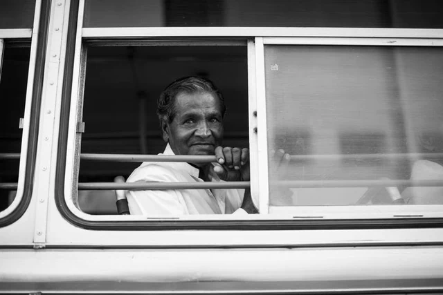 Un passager regarde le photographe par une fenêtre ouverte munie de barres horizontales, en noir et blanc.