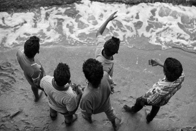 Cinq hommes vus de dessus au bord de l’eau, l’un levant le bras face aux vagues, en noir et blanc.