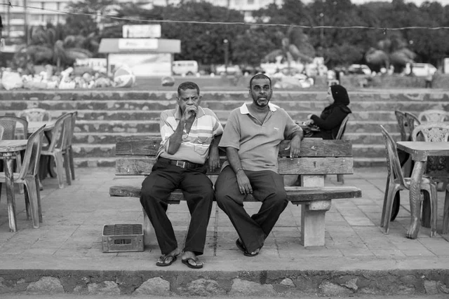 Deux hommes assis sur un banc en bois devant des marches et une terrasse, photographiés en noir et blanc.