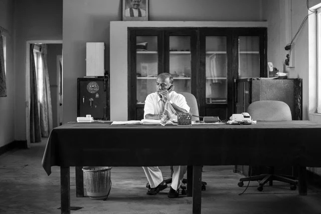 Un homme assis derrière un grand bureau parle au téléphone, en noir et blanc.