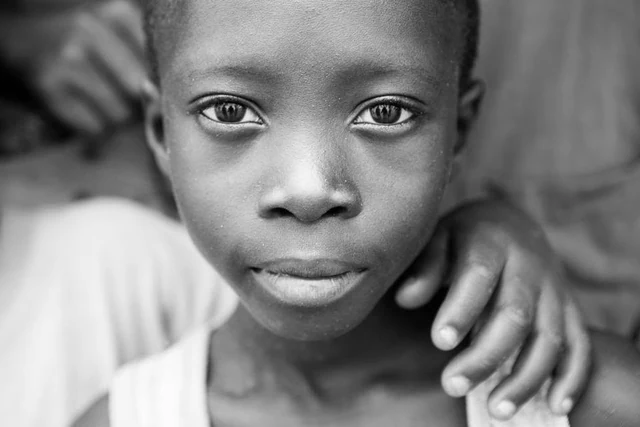 Portrait de face en noir et blanc d’un enfant regardant l’objectif, une main posée sur son épaule.