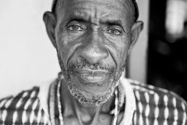 Portrait de face en noir et blanc d’un homme à la barbe courte grisonnante, vêtu d’une chemise à motifs.