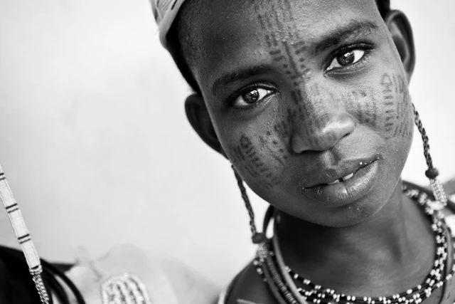 Portrait rapproché en noir et blanc d’une personne portant des colliers de perles, avec des motifs sur le visage et le regard tourné vers l’objectif.