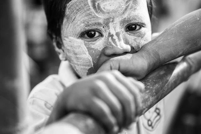 Portrait en noir et blanc d’un enfant au visage couvert de motifs clairs, les bras appuyés sur une barre.