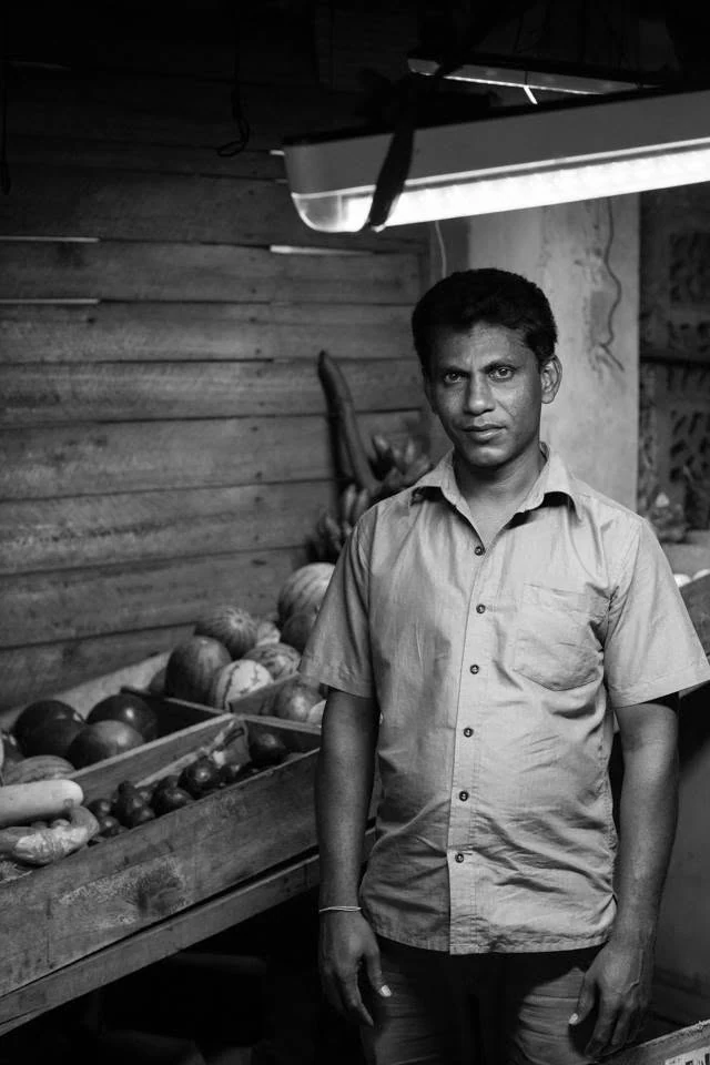 Portrait en noir et blanc d’un homme debout à côté de cagettes de légumes dans une échoppe.
