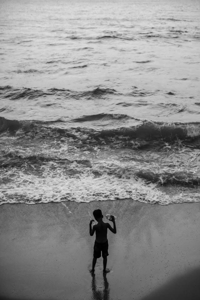 Un enfant vu de dos sur le sable face aux vagues, photographié en plongée et en noir et blanc.