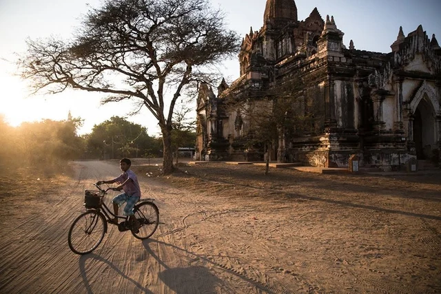 Un enfant à vélo passe devant un temple et un grand arbre, dans une lumière dorée qui projette de longues ombres.