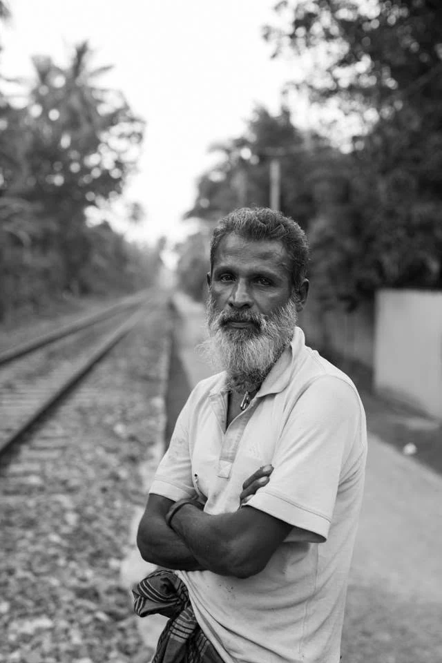 Portrait en noir et blanc d’un homme à la barbe blanche, les bras croisés près d’une voie ferrée.