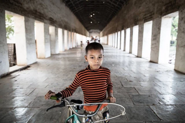 Un enfant en pull rayé rouge et noir tient un vélo dans une longue galerie bordée de colonnes.