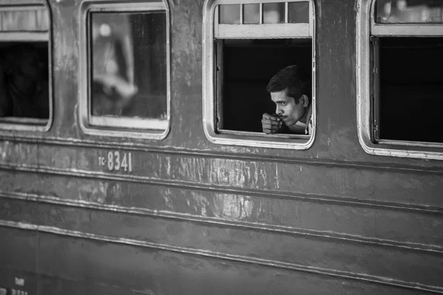 Un homme regarde dehors par la fenêtre ouverte d’un wagon ancien, en noir et blanc.