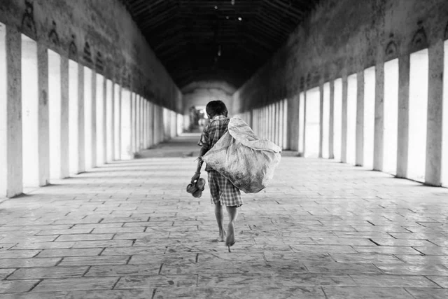 Une personne marche pieds nus dans une longue galerie à colonnes, portant un grand sac et ses chaussures à la main, en noir et blanc.