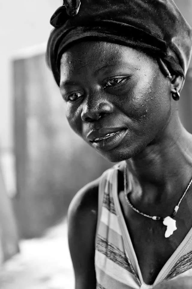 Portrait en noir et blanc d’une femme portant un foulard sombre et un pendentif en forme de continent africain, le regard baissé.