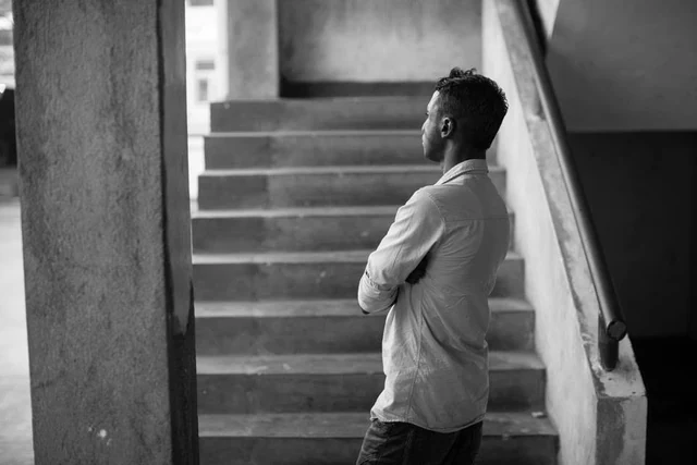 Un homme debout, les bras croisés, au pied d’un escalier, photographié de profil en noir et blanc.