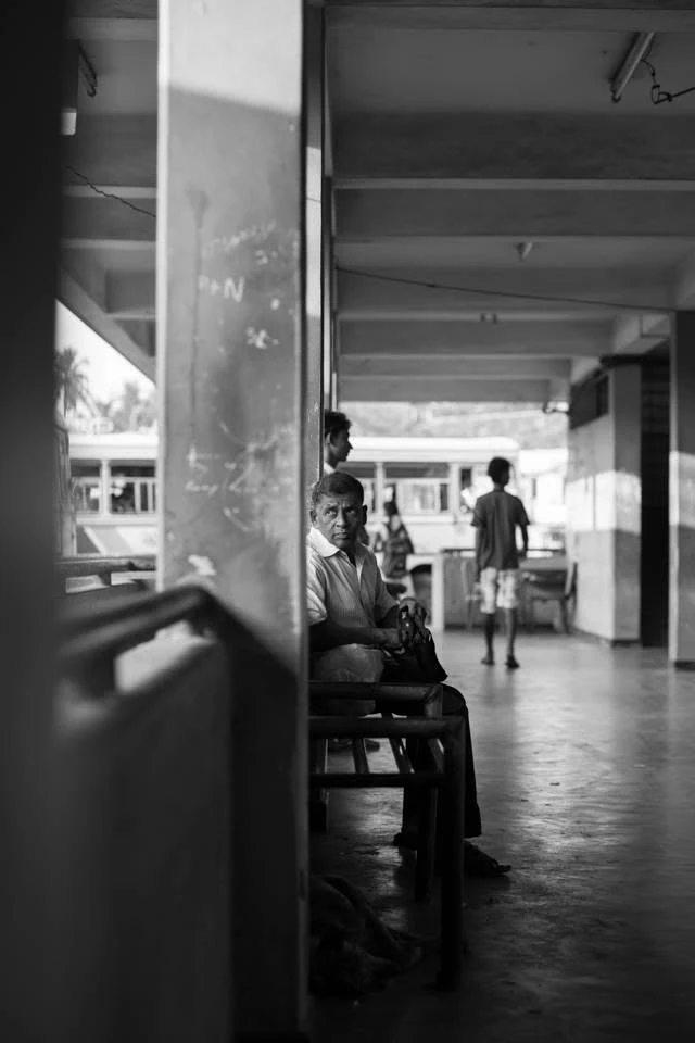 Un homme assis sur un banc entre des piliers regarde le photographe, tandis que des passants s’éloignent, en noir et blanc.