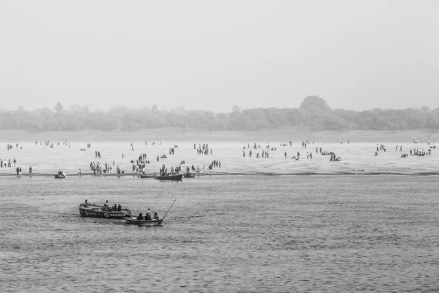 Des barques sur un fleuve et de nombreuses personnes sur la rive opposée, photographiées en noir et blanc en Inde.