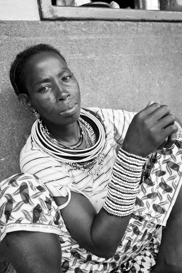 Une femme assise contre un mur porte de nombreux colliers et bracelets de perles, les mains réunies devant elle, en noir et blanc.