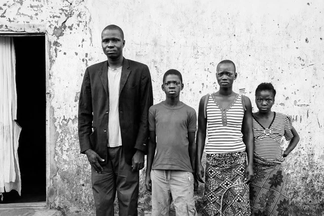 Quatre personnes debout côte à côte devant un mur à la peinture écaillée et une porte ouverte, photographiées de face en noir et blanc.