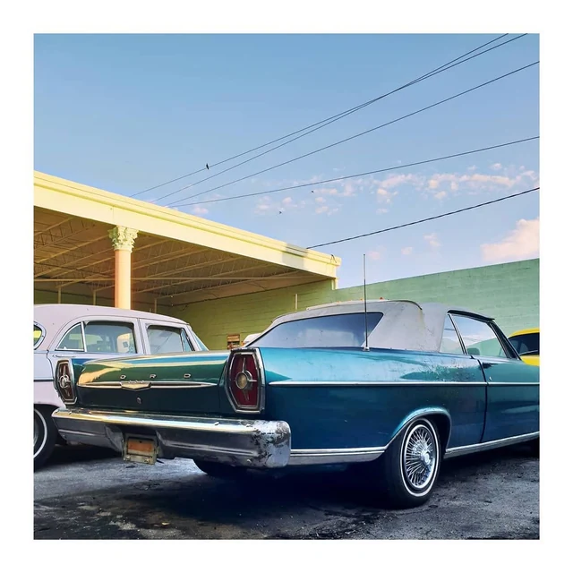 Une voiture ancienne bleu turquoise et blanche devant un garage aux murs pastel, sous un ciel traversé de câbles.