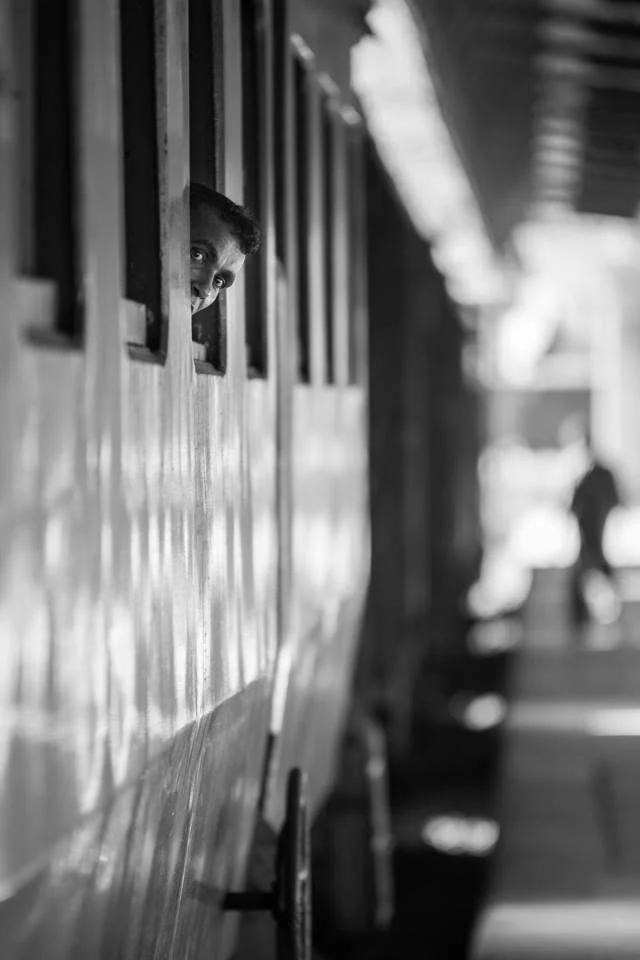 Un visage apparaît à la fenêtre d’un train le long d’un quai, en noir et blanc et faible profondeur de champ.