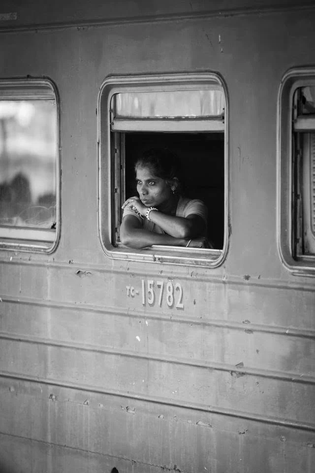 Une femme appuyée sur le rebord d’une fenêtre de train regarde au loin, en noir et blanc.